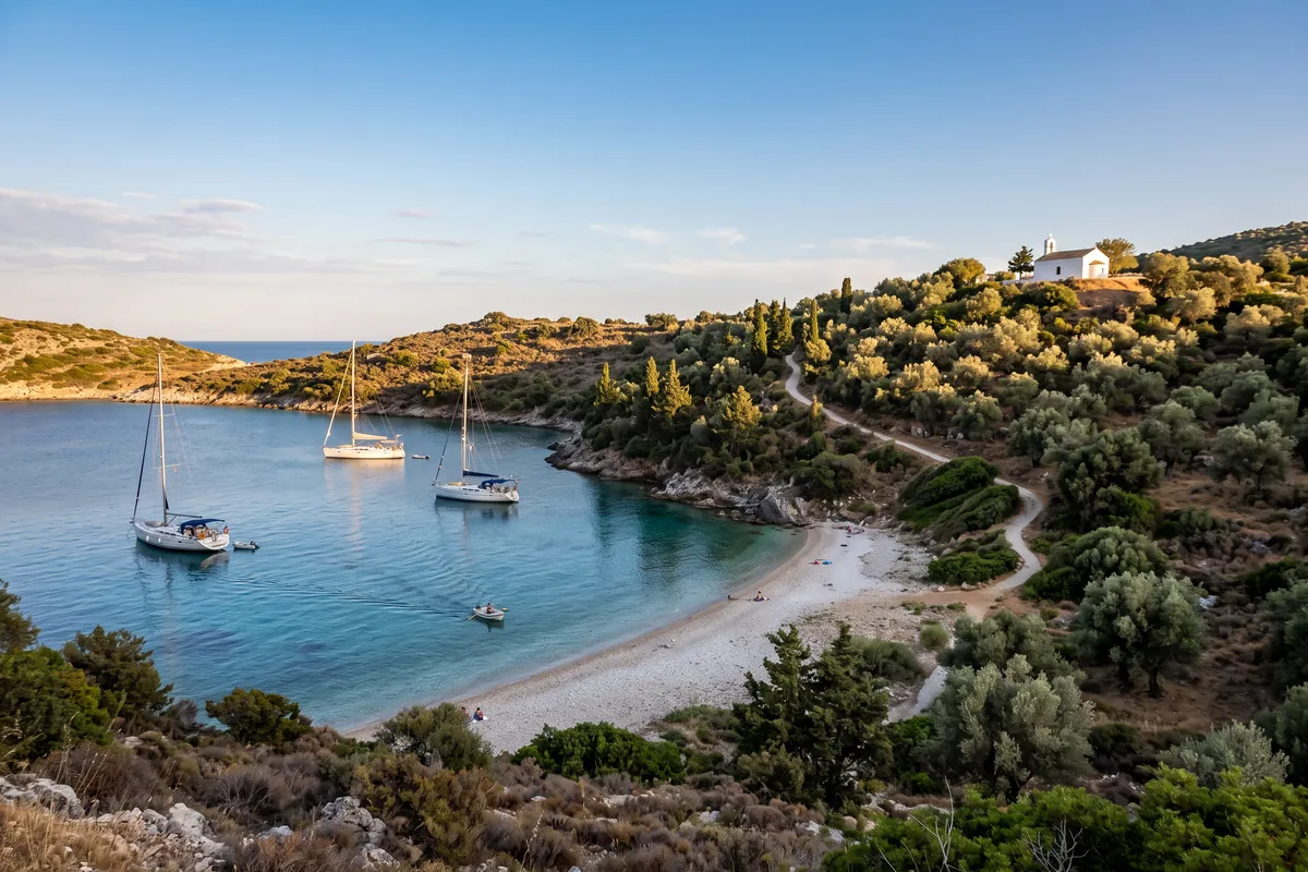Drei Segelyachten ankern in einer Bucht auf einer griechischen Insel, ein Beiboot fährt zum Kiesstrand, ein Pfad führt zu einer weißen Kapelle