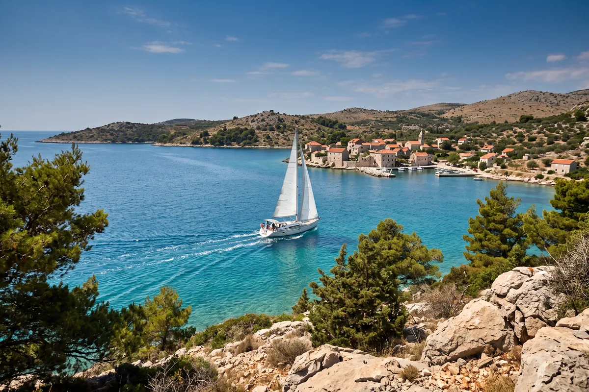 Segelyacht mit weißen Segeln in einer türkisen Bucht an der dalmatinischen Küste, das Kielwasser führt zu einem Steindorf mit Hafen
